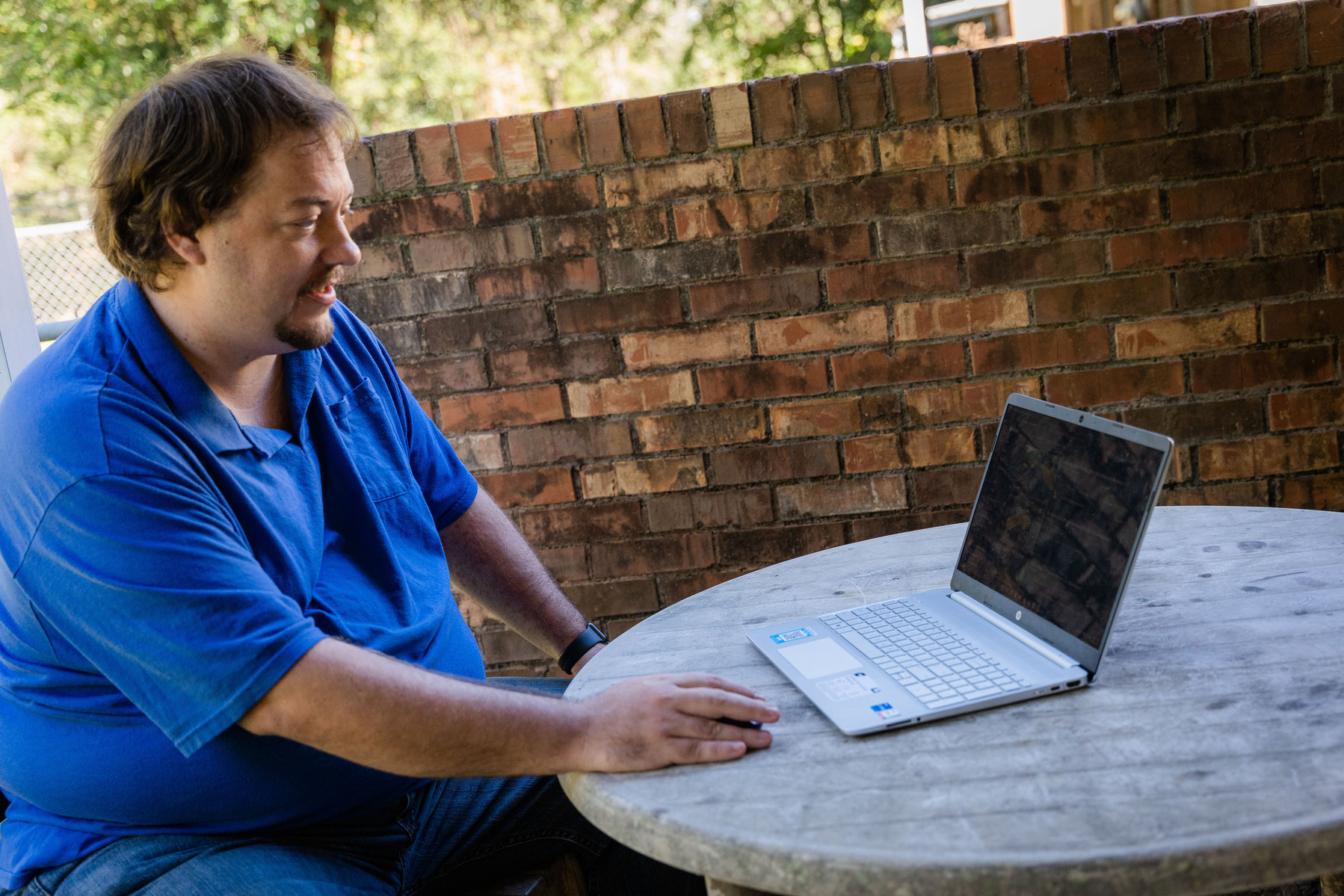 A man in a blue polo sits at a table and works on a laptop computer.