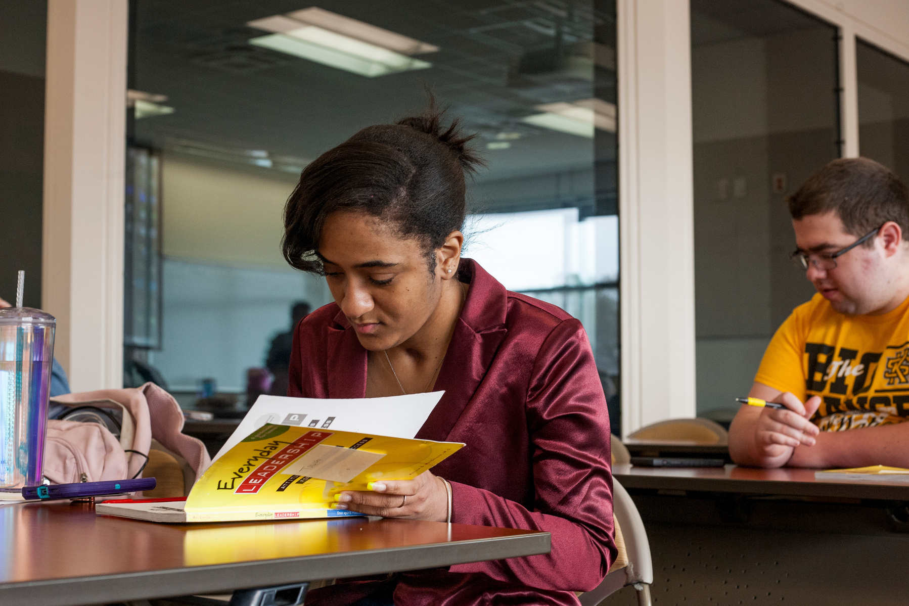 African American woman wears a rose jacket as she works in a yellow text book during class. 