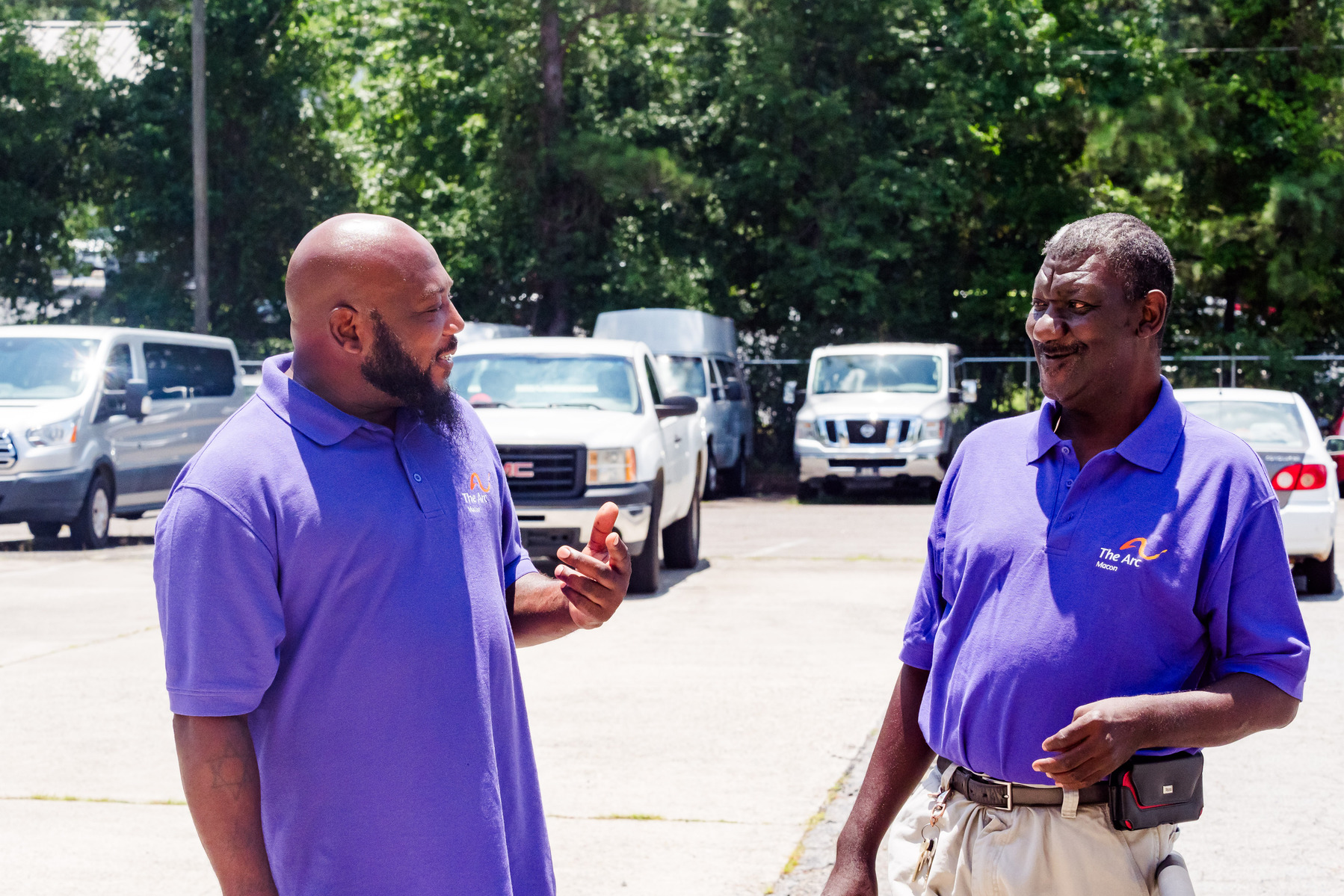 Two African American men wear matching purple polo shirts as they stand and talk in a parking lot. 