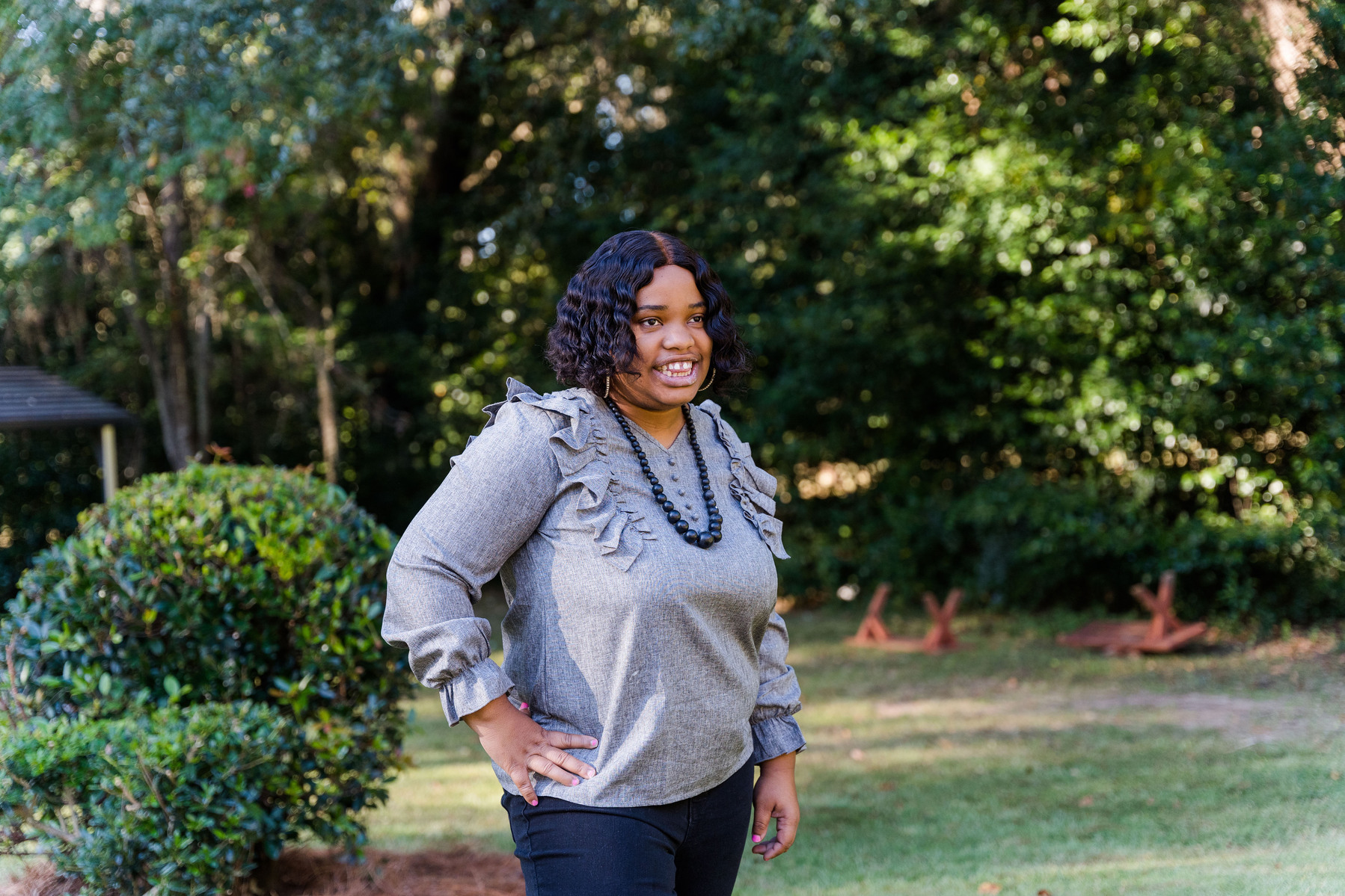 A young woman with dark hair wears a grey blouse and a black necklace. She is standing outside in front of trees and is smiling. 