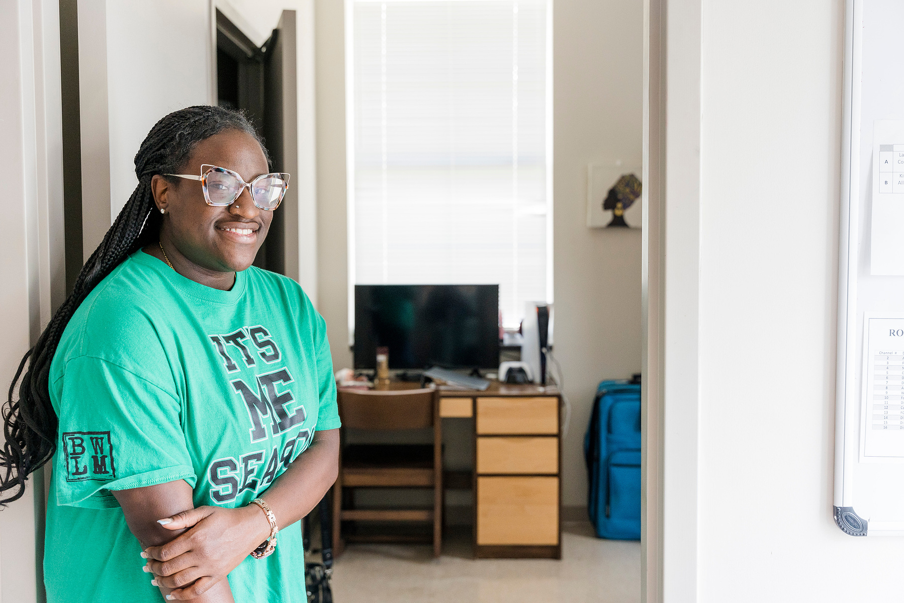 Young African American woman wears braids and a green shirt while standing in a dormitory hallway.