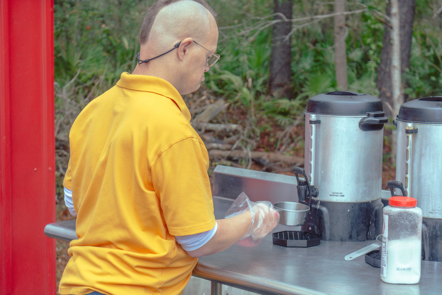 Man in yellow shirt pours a cup of coffee from a steel coffee pot.