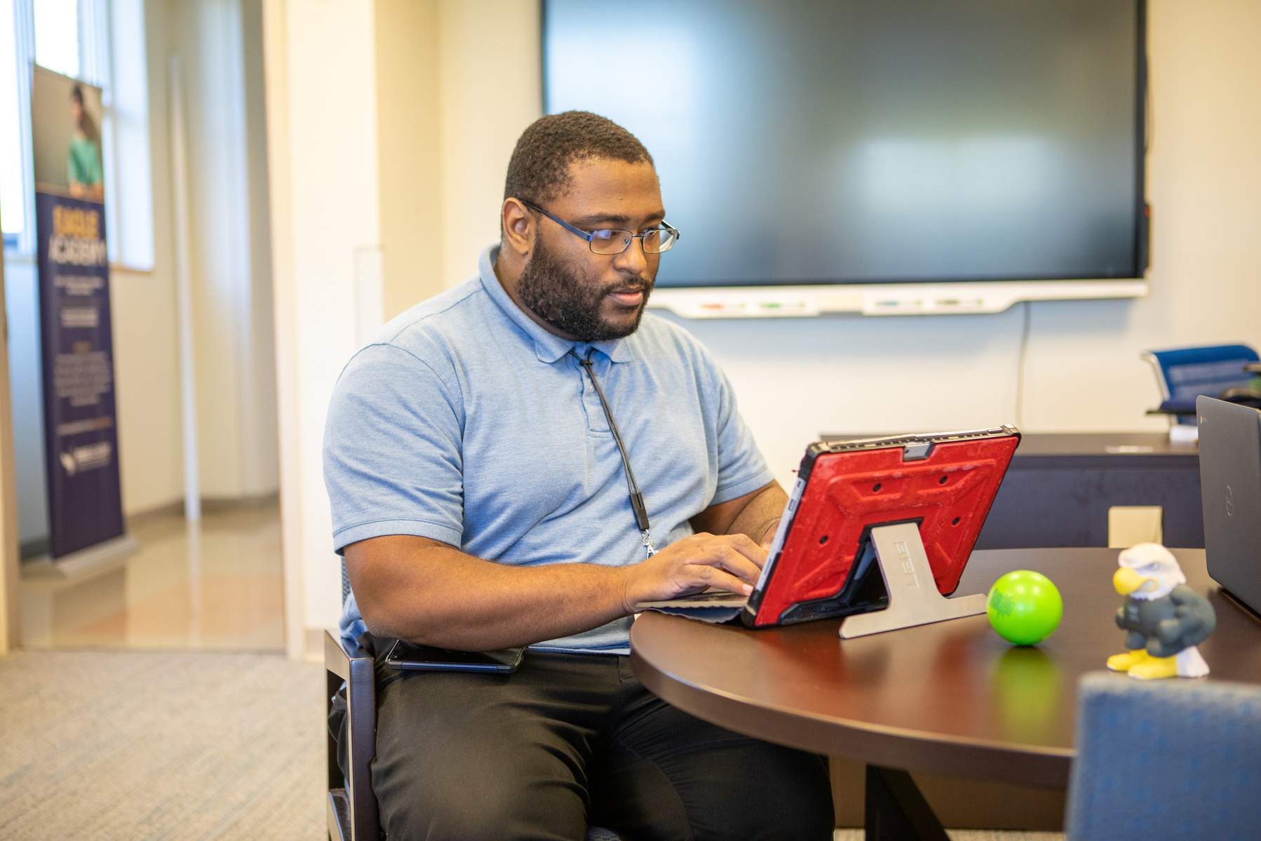 A young black male wears a blue shirt and glasses as he sits at a table and works on a computer. 