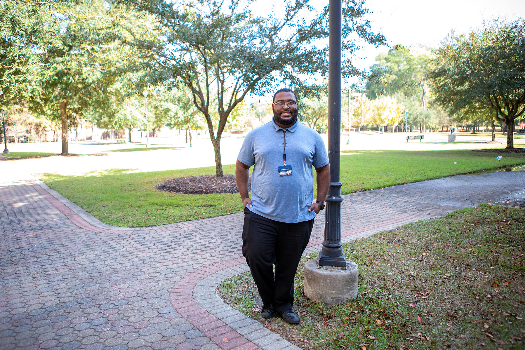 A young black male wears glasses and a blue shirt and stands on a tree-lined path.
