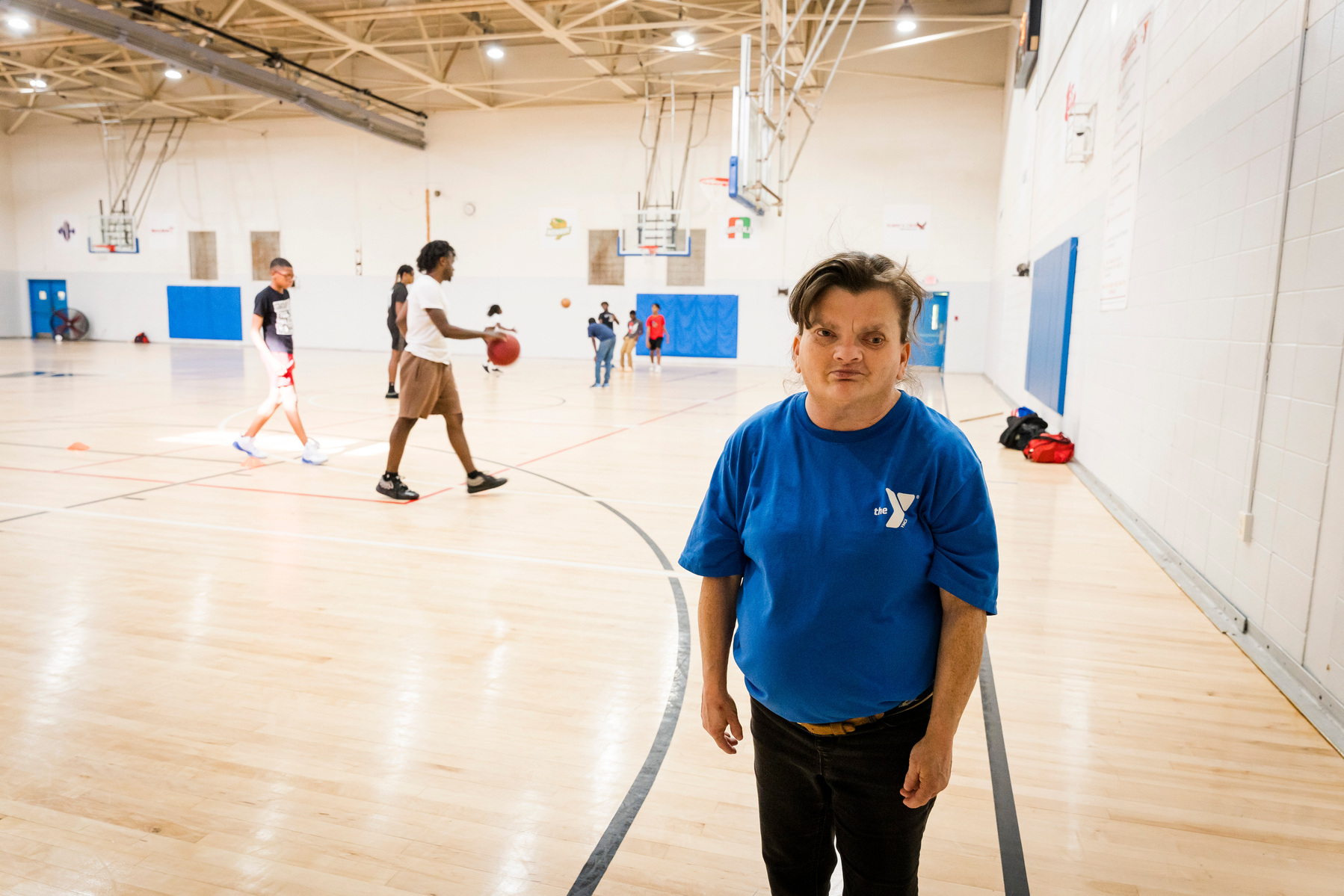 A woman with brown hair stands inside a gymnasium with basketball players on the court. 