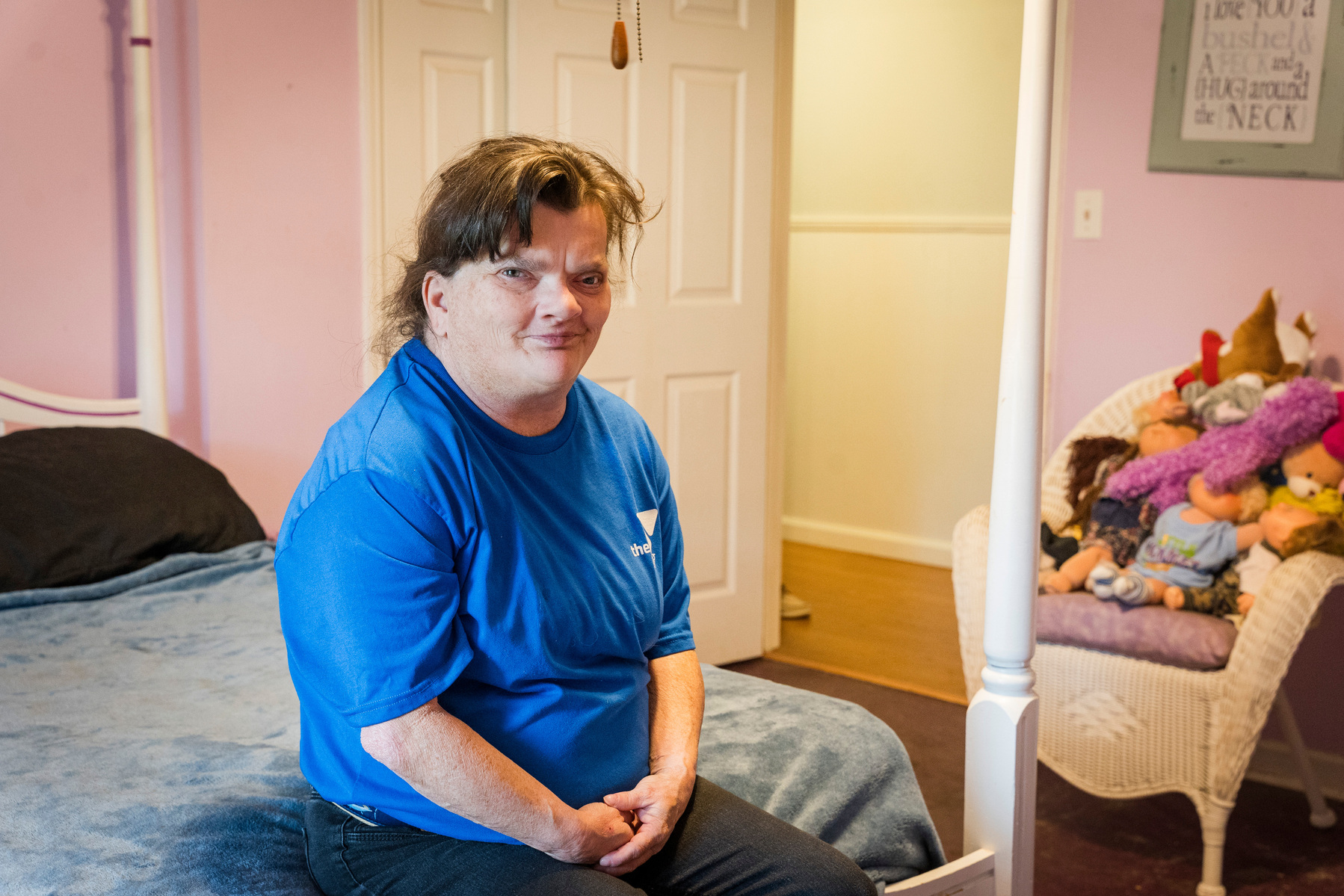 A woman with brown hair wears a blue shirt and sits on the end of a bed.