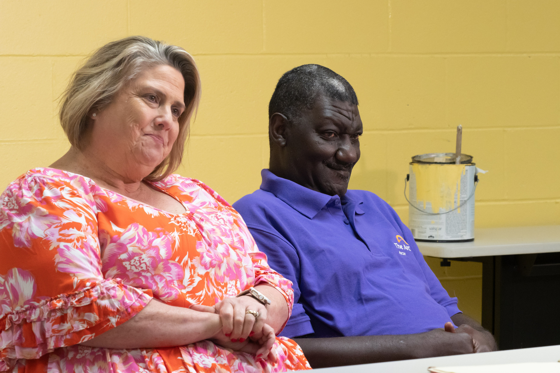A man and a woman wear bright shirts and sit at a table in front of a yellow wall. 