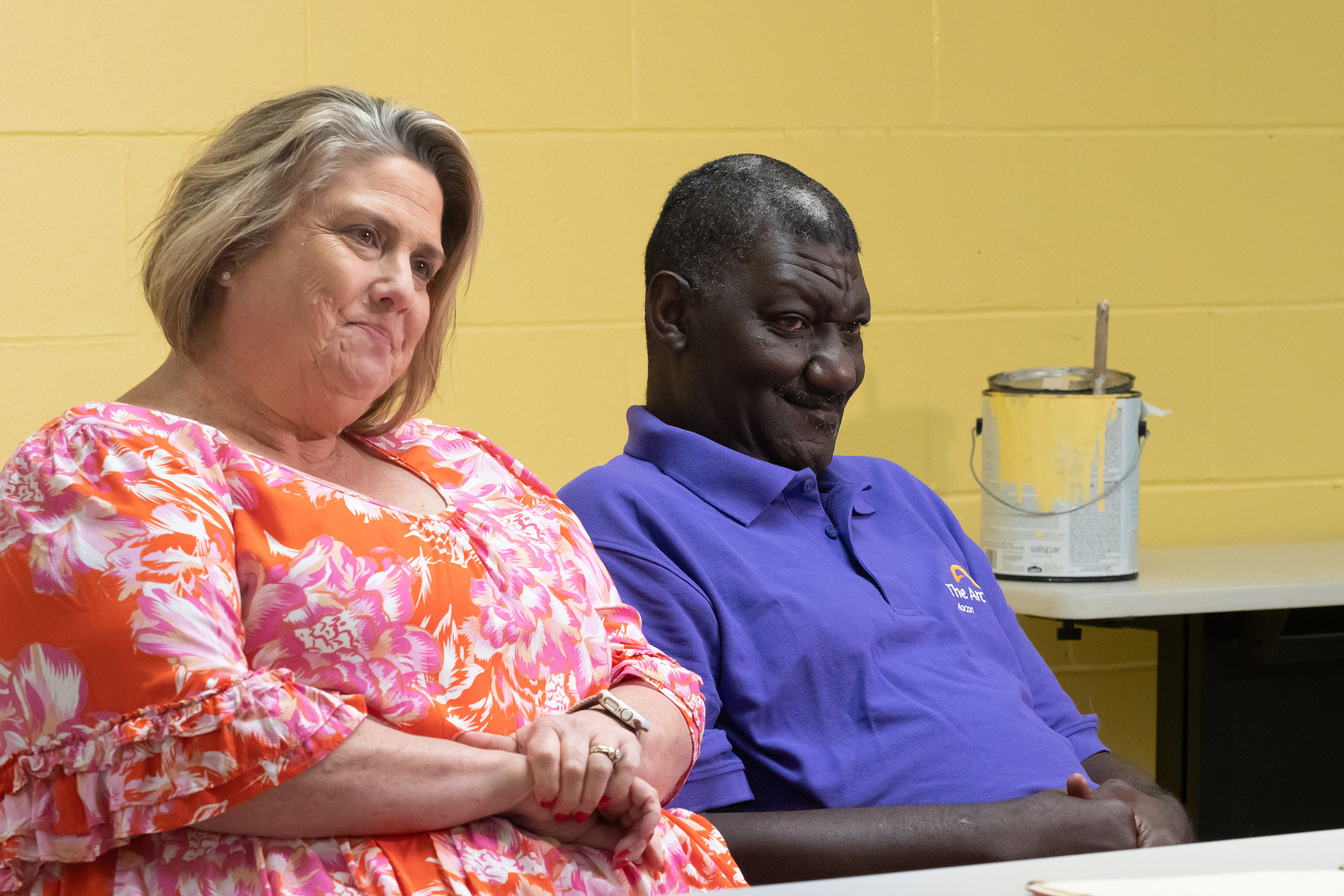 A man and a woman wear bright shirts and sit at a table in front of a yellow wall. 