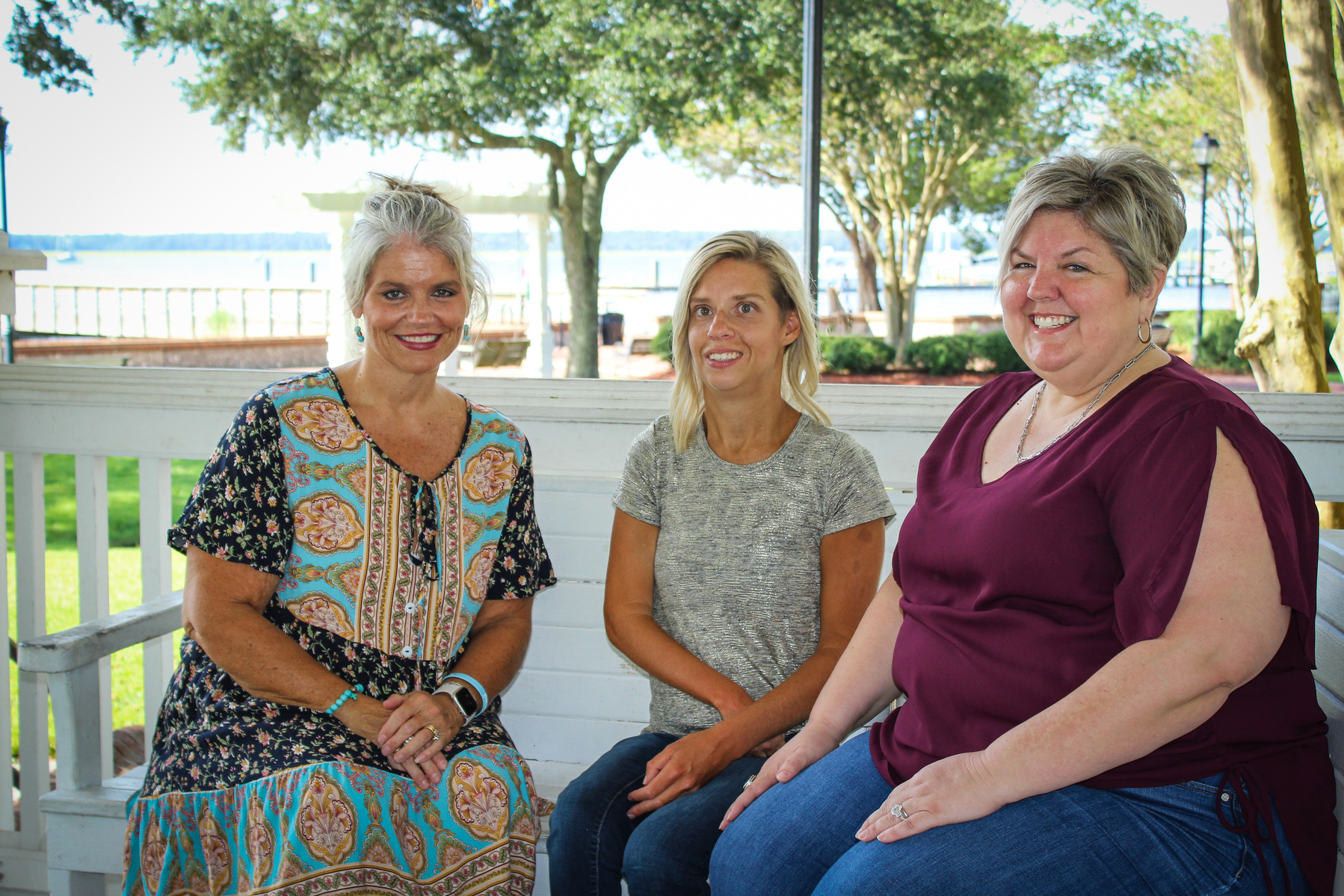 Three women sit on a bench outside and smile for the camera.