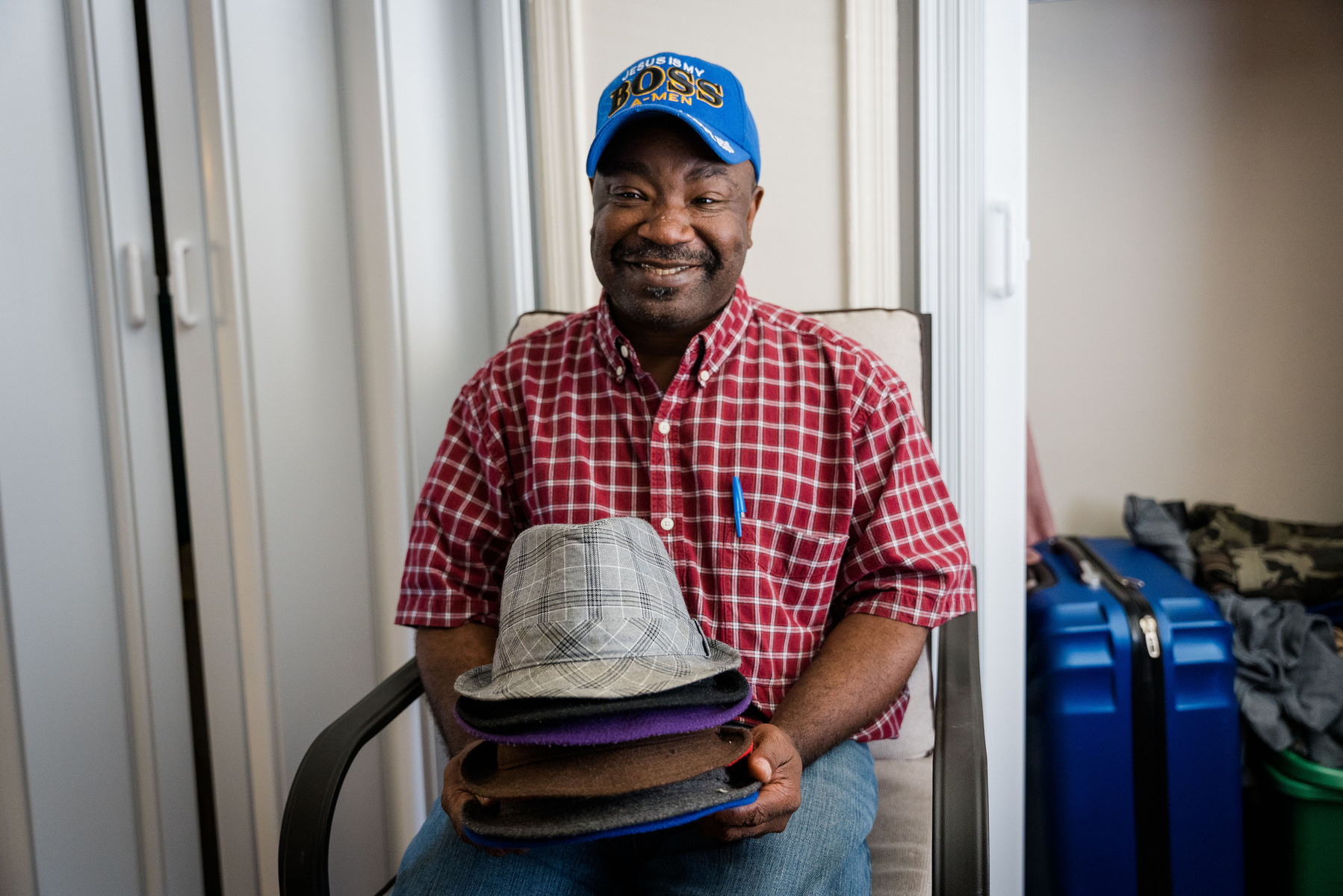 African American man in a plaid shirt and blue ball cap smiles in his living room while holding a stack of hats.