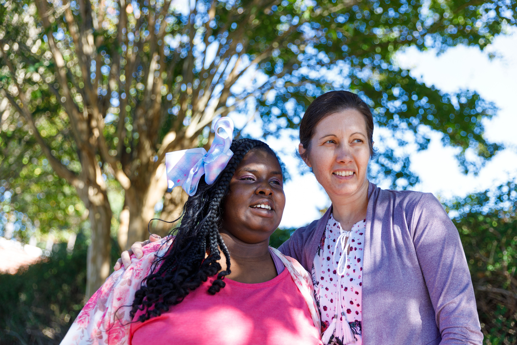 Mother and daughter stand outside under a tree on a sunny day. 