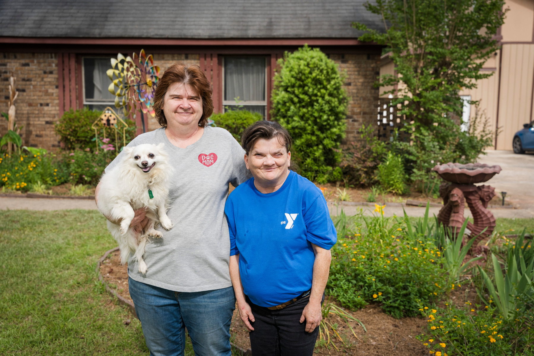 Two women stand in front of a house as one of them holds a dog. 