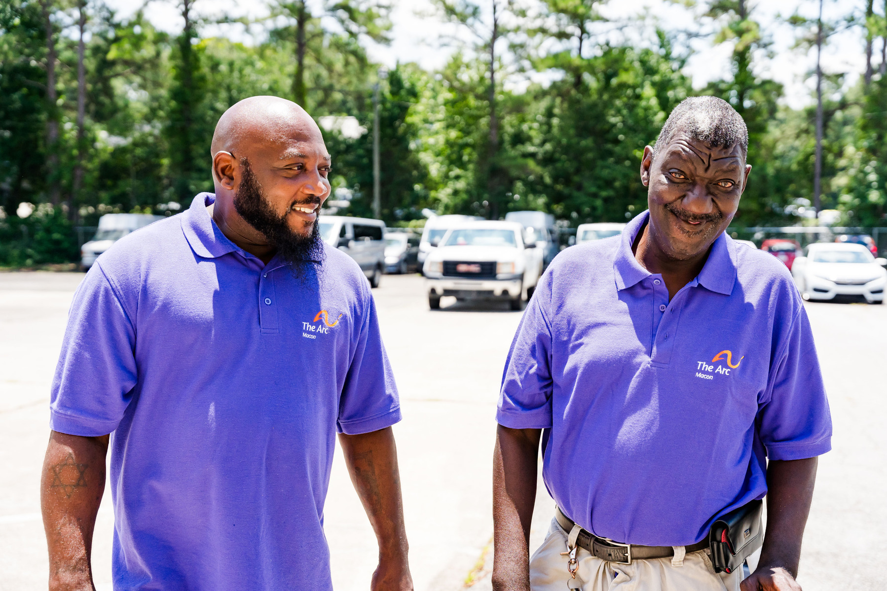 Two African American men wear matching purple polo shirts as they stand and talk in a parking lot. 