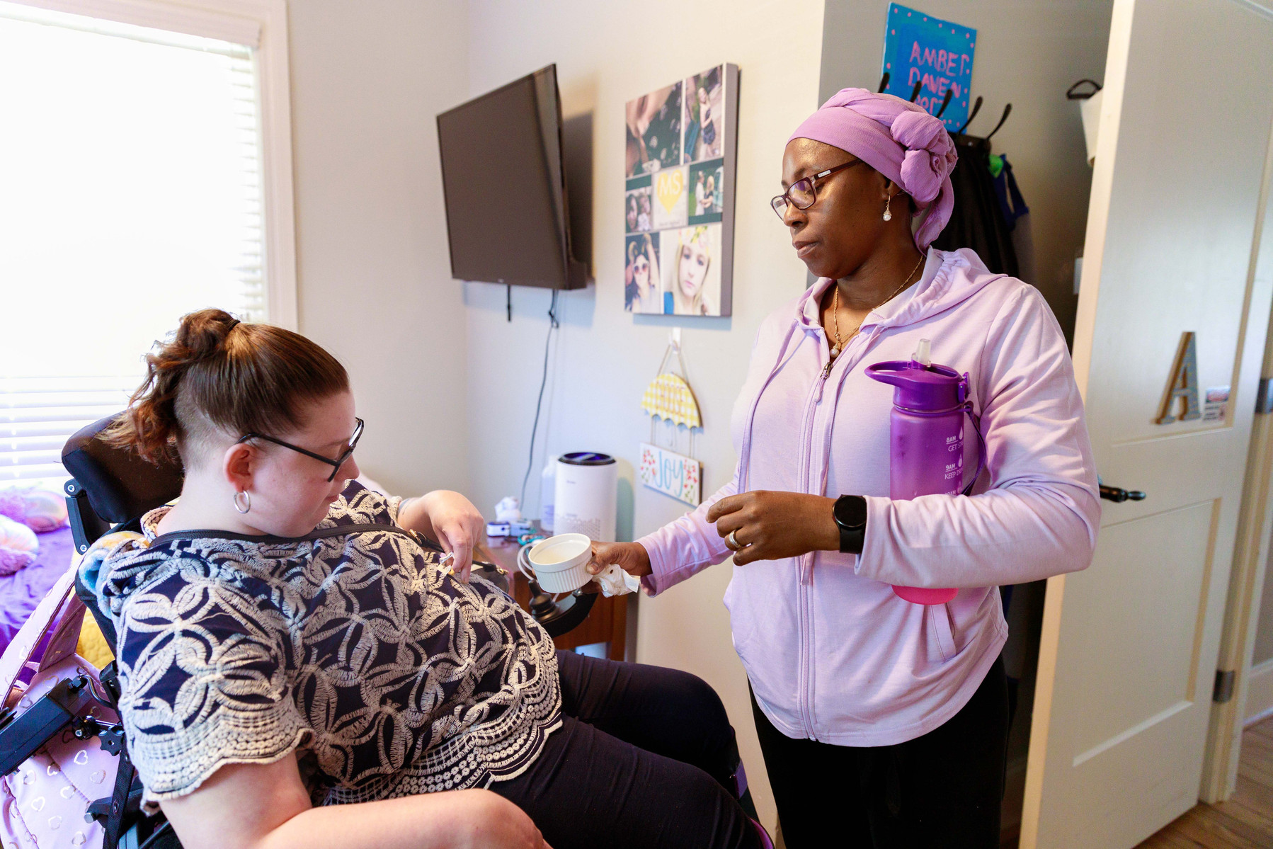 A young brunette woman in a patterned shirt and glasses takes medication from a care attendant wearing a pink jacket and headwrap.