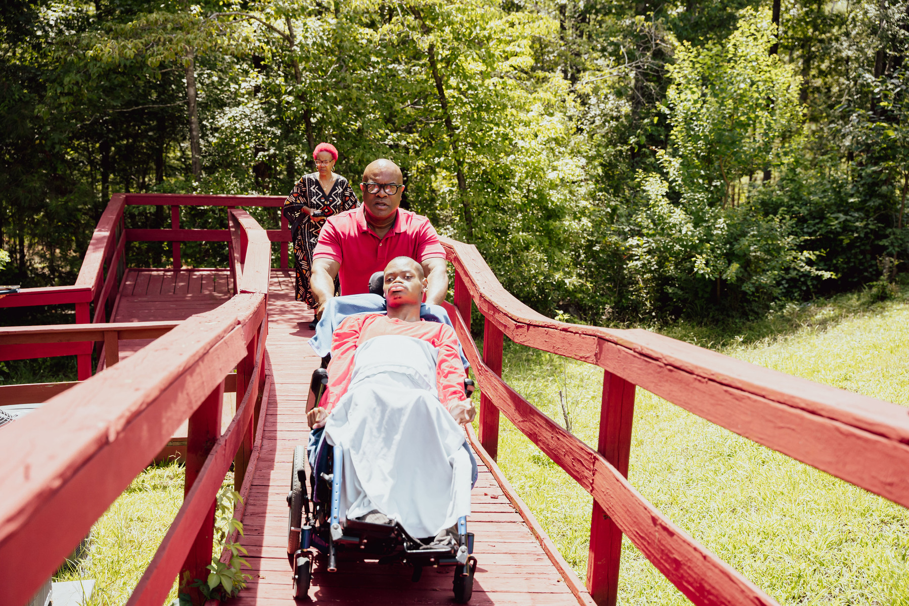 A father pushes his son in a wheelchair as his wife follows them on a red walkway. 
