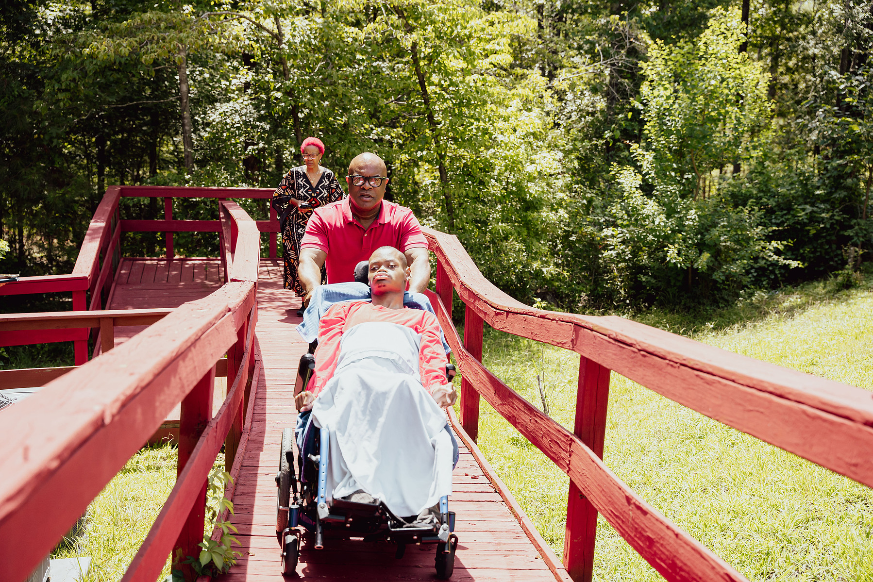 A father pushes his son in a wheelchair as his wife follows them on a red walkway. 