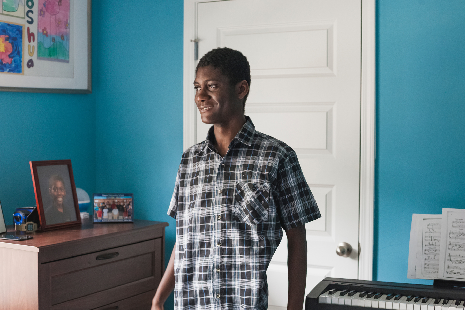 A young African American man in a plaid shirt stands in his blue bedroom. 
