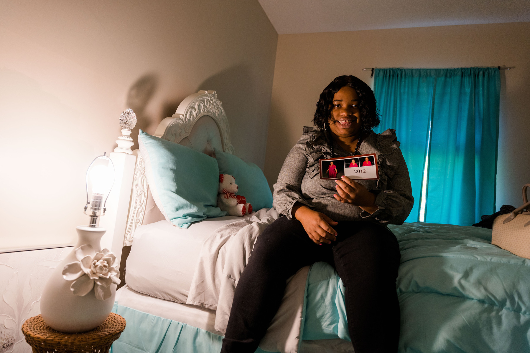 A young woman in a grey blouse sits on a pastel-colored bed in a bedroom.