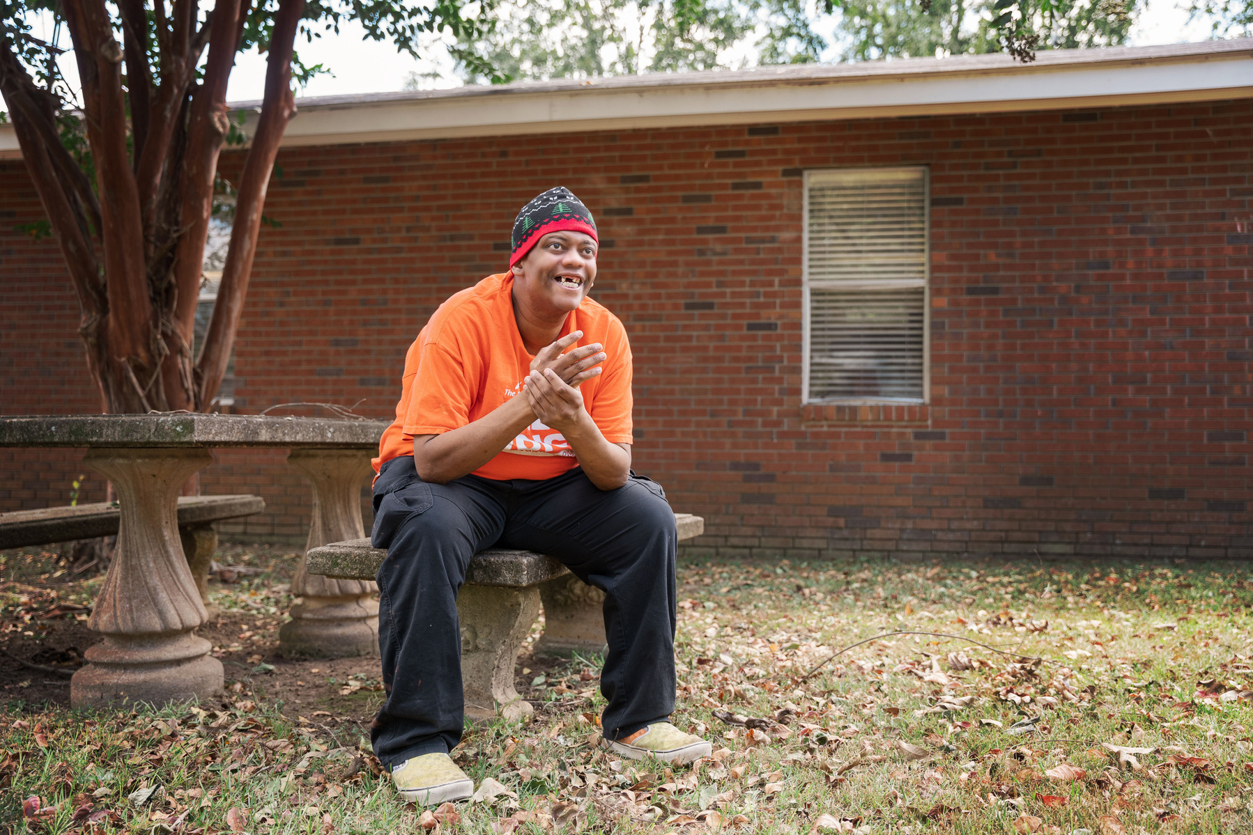 A man in an orange shirt and knit cap sits outside on a bench smiling.