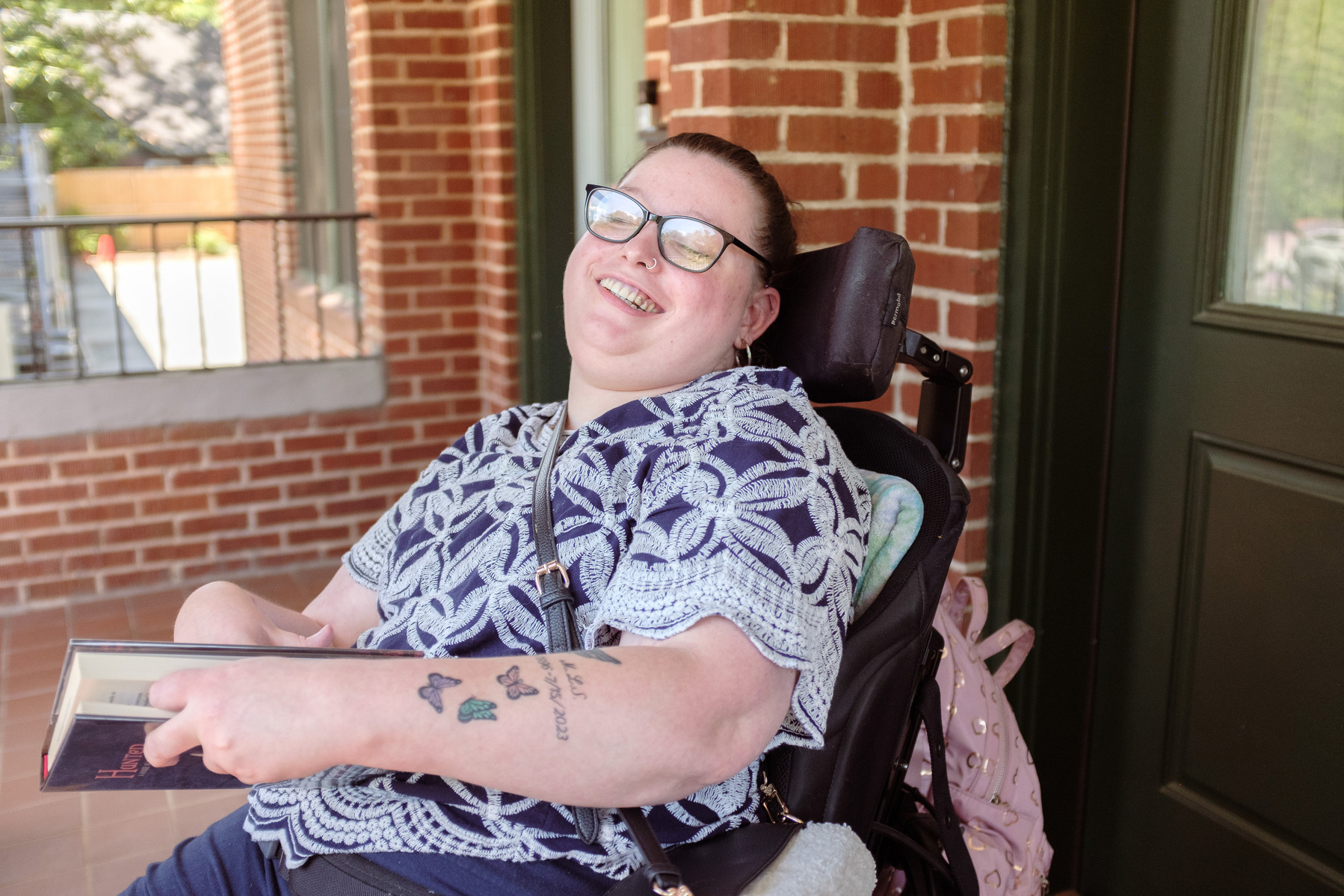 A young brunette woman in a patterned shirt and glasses holds a book and sits on a screened porch. 