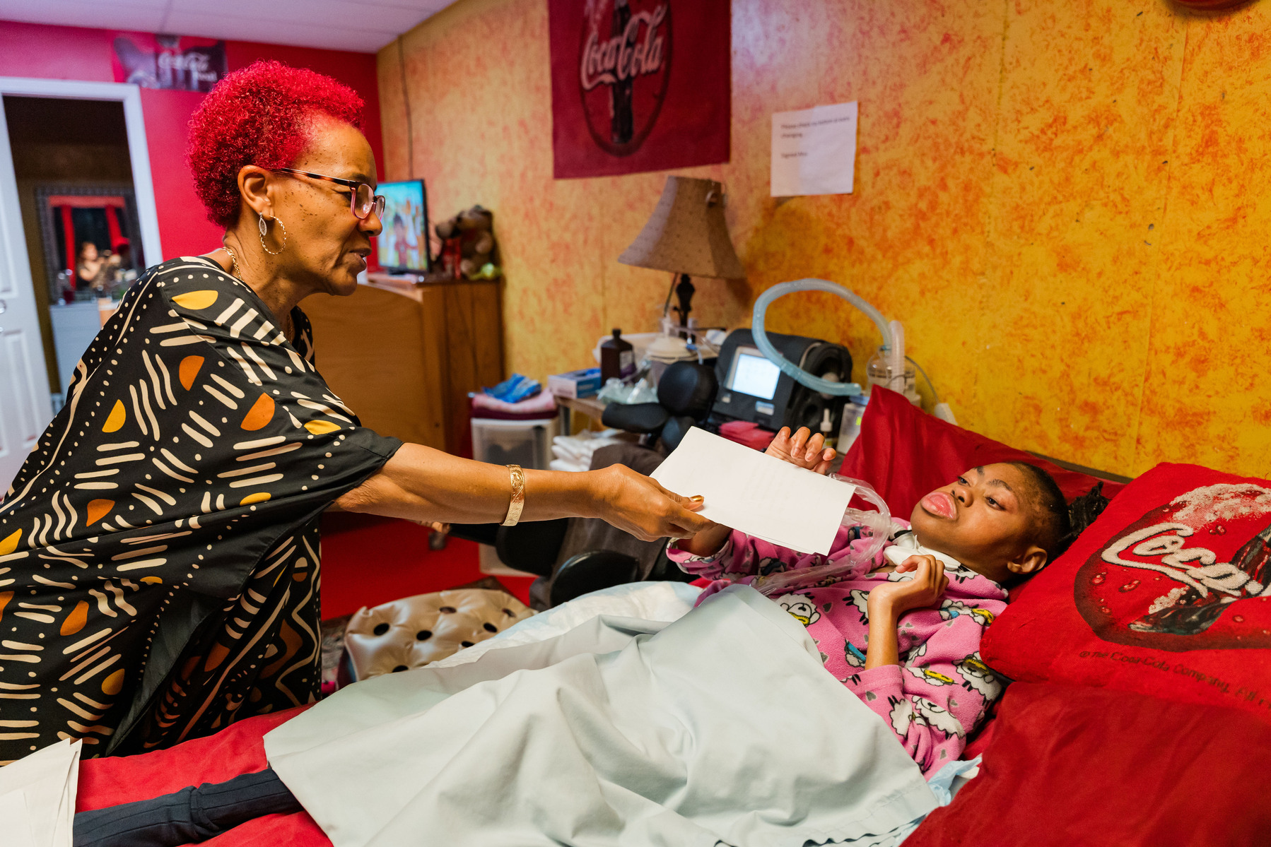 A woman with red hair hands her daughter a sheet of paper as she lies in bed. 