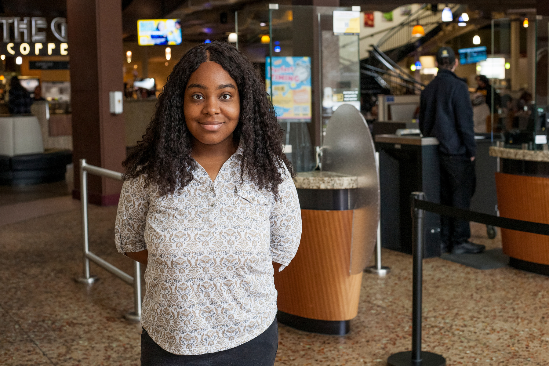 A young African American woman in a white blouse stands in a college cafeteria. 
