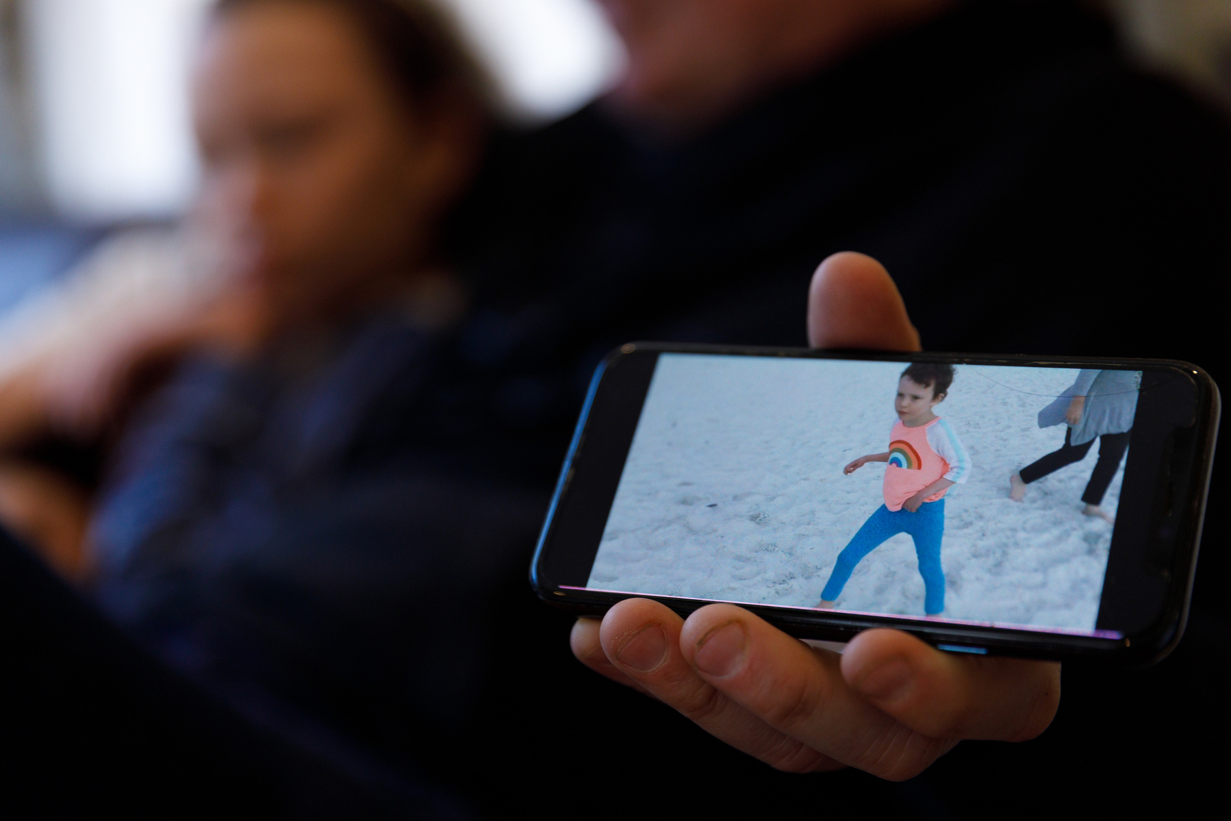 A phone shows a photo of a toddler running on a beach. The same girl, now 11, sits behind the phone. 