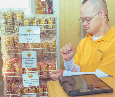 Men wears glasses and a yellow shirt as he stands in front of a shelf full of popcorn.