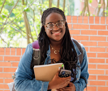 A young African American woman with her hair in braids wears glasses, a denim jackets and holds books. 