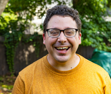 A young man with brown curly hair and glasses smiles for the camera. 