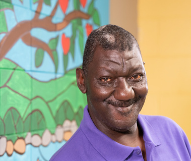 A middle aged African American man wears a purple polo shirt and paints a wall cream.