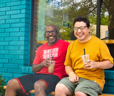 Two men sit on a bench in front of a blue brick store and smile. 