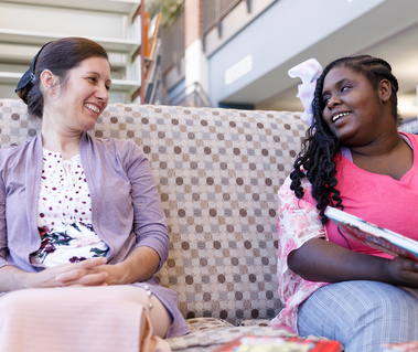 Mother and daughter sit on a patterned sofa and smile at each other. 