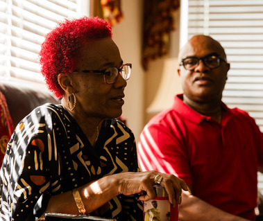 A woman with red hair sits on a sofa next to her husband and talks. 