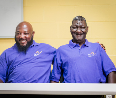Two middle aged African American men wear matching purple polos and sit at a table. 