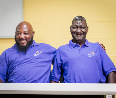 Two middle aged African American men wear matching purple polos and sit at a table. 