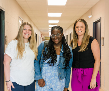 A young African American woman with her hair in braids stands in between two women in a hallway.