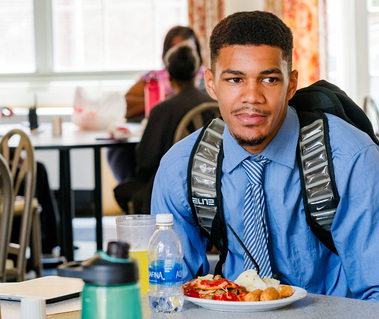 A young African American man wears a blue dress shirt and tie as he dines in a cafeteria.
