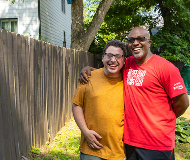 Two men stand outside next to a wooden fence and smile. 