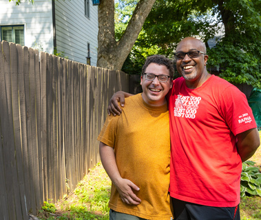 Two men stand outside next to a wooden fence and smile. 