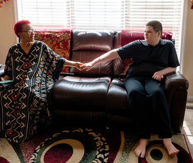 A woman with red hair sits on a sofa and holds hands with her teenage son. 
