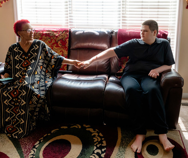 A woman with red hair sits on a sofa and holds hands with her teenage son. 