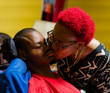 A woman with red hair kisses the cheek of a young man in a wheelchair. 