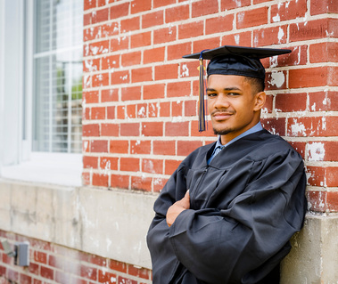 African American young man wears a graduation cap and robe as he leans against a brick building.