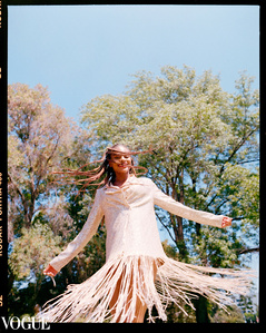 A joyful woman model wearing a shimmering, fringed dress dances under a sunny sky with trees in the background, captured by a fashion photographer. Published in Vogue Italia PhotoVogue.