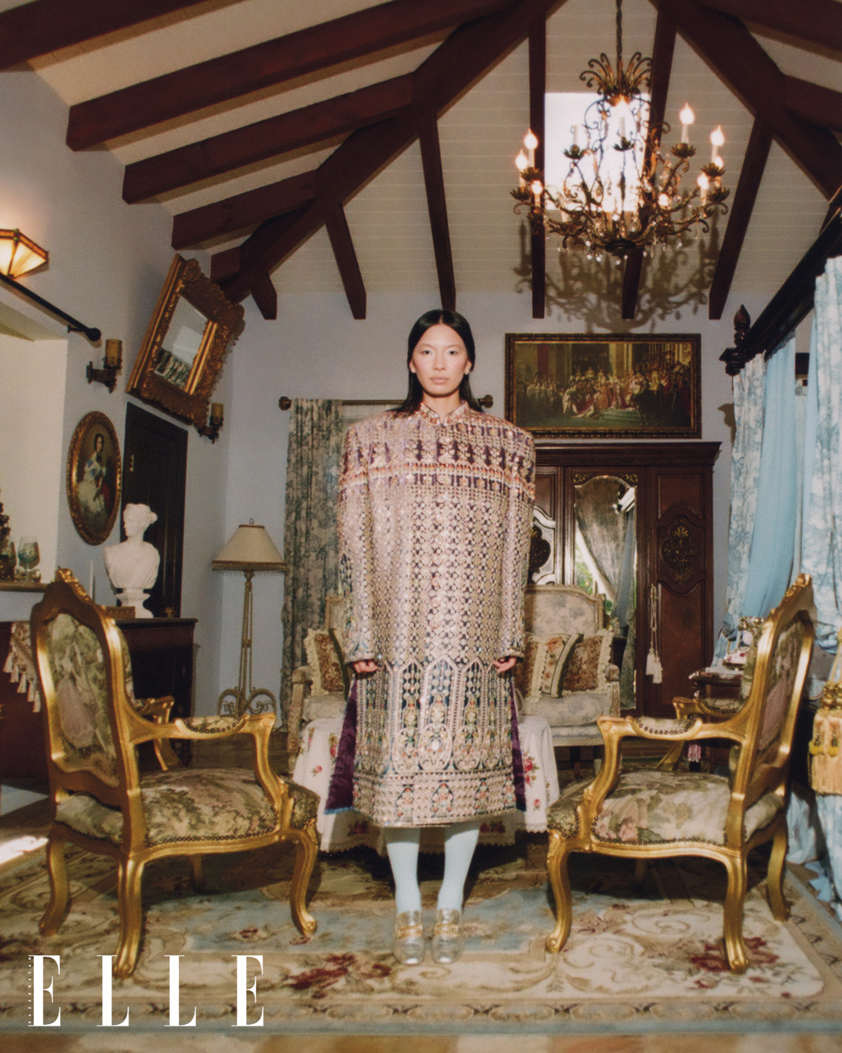 Asian-American female fashion model Mei Li Zheng wearing a custom bejeweled gold Indian Sherwani, baby blue leggings, and silver designer shoes, standing in the center of a Rococo and Baroque style bedroom.