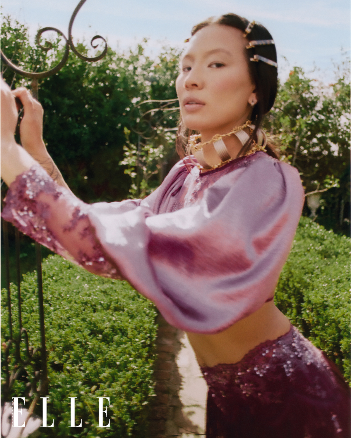 Fashion editorial photograph featuring female Asian American model Mei Li Zheng, outdoors in a lush green garden under blue skies, wearing couture designer jewelry, a purple satin puff sleeve top with lace and crystal applique sleeve, and fringe skirt. 