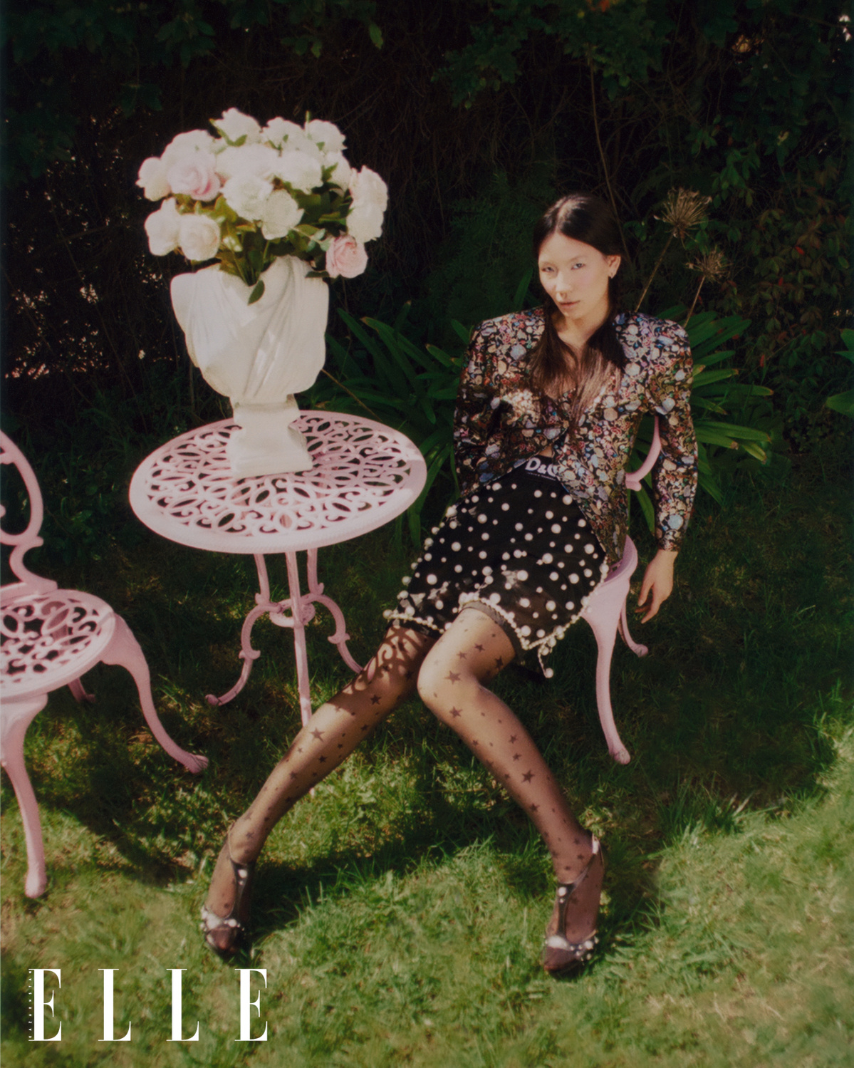 Asian-American female model Mei Li Zheng sits in a pink garden chair, a pot of pink and white flowers on a bust atop a tea table next to her. Mei Li wears a vintage blazer, shorts from Dolce & Gabbana, and black designer shoes from Tinamarie. 