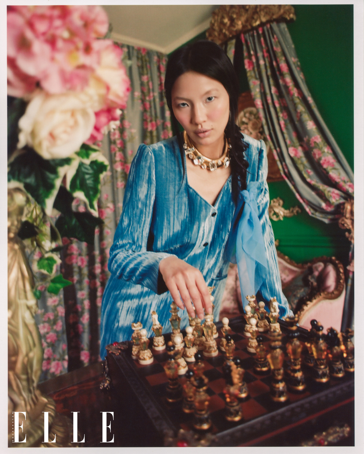 Asian-American female model Mei Li Zheng stares into the camera as she is playing with a chess set, with a pot of flowers in the foreground. Mei Li wears a blue velvet designer jacket, and gold designer necklace with diamonds. Behind her, floral curtains.