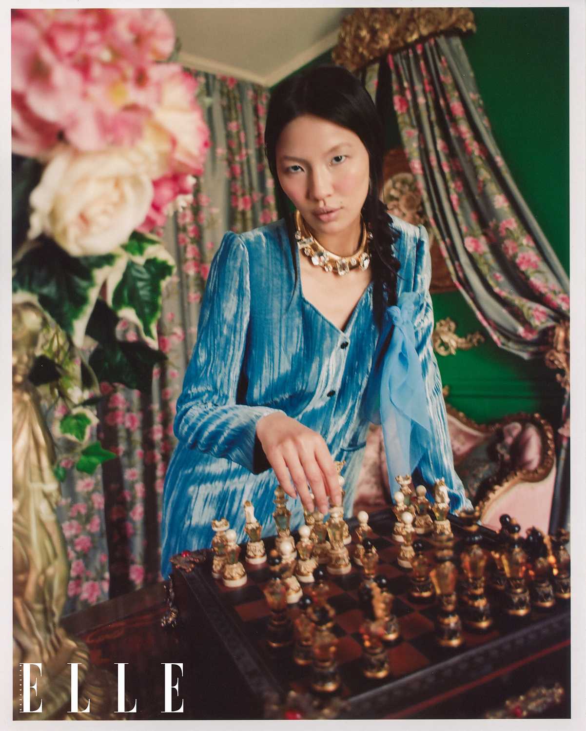 Asian-American female model Mei Li Zheng stares into the camera as she is playing with a chess set, with a pot of flowers in the foreground. Mei Li wears a blue velvet designer jacket, and gold designer necklace with diamonds. Behind her, floral curtains.